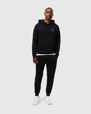 A man is standing against a plain, light grey background, wearing a Psycho Bunny MENS LEVY FLEECE HOODIE - B6H859D200 with a small emblem on the chest, black jogger pants, and white sneakers. His hands are in his hoodie pockets, and he is looking at the camera with a neutral expression.
