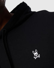 Close-up of a person wearing a Psycho Bunny Men's Levy Fleece Hoodie (B6H859D200) in black French terry, adorned with a white embroidered logo on the chest. The logo showcases a cartoon bunny with crossbones below it. The 100% cotton hoodie is slightly wrinkled, revealing only part of the person's torso and shoulder.