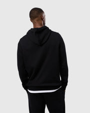 A person stands with their back to the camera, wearing a Psycho Bunny MENS LEVY FLEECE HOODIE - B6H859D200 and black pants. The individual appears to be looking to their left. The background is plain and light gray.