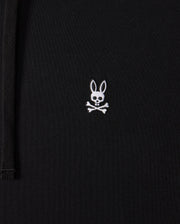 Close-up of a black Mens Levy Fleece Hoodie (B6H859D200) by Psycho Bunny, featuring a small embroidered design of a white bunny head with long ears above two crossed bones, resembling a pirate skull and crossbones. This 100% cotton garment combines comfort with unique style.