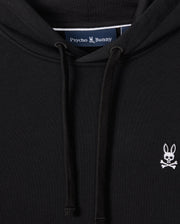 A close-up of the MENS LEVY FLEECE HOODIE - B6H859D200 from Psycho Bunny, featuring a small white embroidered logo of a bunny skull and crossbones on the left chest, showcases its sleek black French terry fabric. The 100% cotton label inside reads "Psycho Bunny," and drawstrings are visible in the foreground, making it an essential piece.