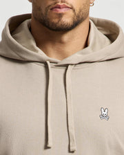 The individual is dressed in a beige Men's Levy Essential Fleece Hoodie (B6H859D200) from Psycho Bunny, made from premium 100% cotton fleece and featuring a small embroidered logo of a bunny with crossed bones on the chest, highlighting its distinguished design details and high-quality fabric.