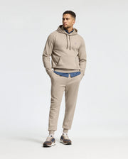 A man casually stands with his hands in his pockets, sporting a Psycho Bunny Men's Levy Essential Fleece Hoodie (100% cotton) and matching sweatpants over a denim shirt. He's wearing sneakers while looking slightly to the side against a plain background, adding a touch of comfort to his laid-back style.