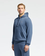 A person stands wearing a Psycho Bunny MENS LEVY ESSENTIAL FLEECE HOODIE in blue, paired with matching pants against a plain light background. This 100% cotton sweatshirt features a small embroidered logo on the left chest, embodying modern men's fashion.