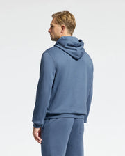 A person with short blond hair faces away from the camera, wearing Psycho Bunny's Men's Levy Essential Fleece Hoodie and matching pants in soft blue. Crafted from 100% cotton, the ensemble is set against a plain, light-colored background.