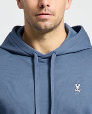 A person in a Psycho Bunny MENS LEVY ESSENTIAL FLEECE HOODIE with a bunny emblem on the chest. The cropped shot highlights men's fashion, showing only the lower face and torso. Made of 100% cotton, this blue hoodie features drawstrings and a smooth texture.