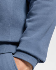 A person wearing a Psycho Bunny MENS LEVY ESSENTIAL FLEECE HOODIE (B6H859D200) showcases the 100% cotton blue sweatshirt with ribbed cuffs. Their hand is partially visible inside the pocket of matching blue sweatpants, highlighting the soft and comfortable fabric.
