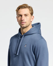 A short-haired blond person wears a Psycho Bunny Men's Levy Essential Fleece Hoodie (B6H859D200) in blue, with a small embroidered rabbit logo on the chest. Made from 100% cotton, this hoodie is a men's fashion staple. The background is plain white.