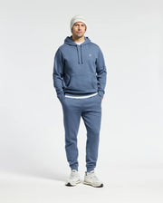 A person dressed in a Psycho Bunny MENS LEVY ESSENTIAL FLEECE HOODIE, joggers, white sneakers, and a white knit beanie stands with hands in pockets against a plain background, embodying men's fashion.