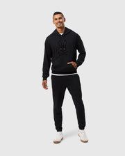 A man wearing the Psycho Bunny MENS PACIFIC CHENILLE HOODIE - B6H930D200, crafted from black, 100% cotton French terry, along with matching jogger pants, stands against a plain light gray background. He smiles with one hand in his hoodie pocket, showcasing the distinctive chenille embroidery patch. He completes the look with white sneakers.