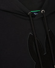 Close-up of the Psycho Bunny Men's Pacific Chenille Hoodie (B6H930D200) in black French terry, showcasing a textured chenille embroidery patch of bunny ears on the front. This 100% cotton hoodie includes drawstrings with metal eyelets, and a small portion of the blue inner tag can be seen at the top.