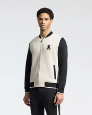 A man showcases effortless style in the Psycho Bunny MENS CRETE COLOR BLOCK BOMBER - B6J421F200, a black and white varsity jacket with classic cut and chest embroidery, paired with black pants against a white background.