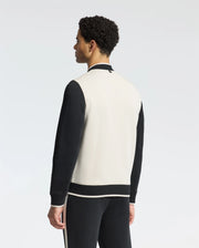 A person with curly hair stands facing away, wearing Psycho Bunny's classic MENS CRETE COLOR BLOCK BOMBER - B6J421F200 in black and white, paired with matching track pants, set against a plain white background.