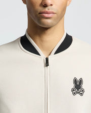 A person models the MENS CRETE COLOR BLOCK BOMBER - B6J421F200 by Psycho Bunny, showcasing its classic cut in beige with a black collar and a bunny head over crossbones emblem on the left side against a plain background.
