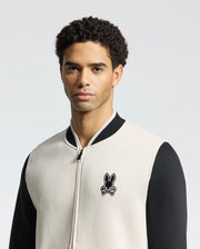 A curly-haired man wears the Psycho Bunny MENS CRETE COLOR BLOCK BOMBER - B6J421F200 in cream and black, featuring a bunny skull logo. He stands against a plain backdrop, looking at the camera, showcasing an ideal transitional outfit.
