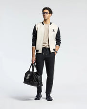 Man in glasses sports a classic look with the Psycho Bunny MENS CRETE COLOR BLOCK BOMBER jacket over a beige shirt, black pants, and shoes. He carries a logo-adorned black duffel bag on his left—a perfect transitional outfit against the white background.