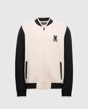 The Psycho Bunny MENS CRETE COLOR BLOCK BOMBER - B6J421F200 is a varsity-style jacket with a beige front and black sleeves. It includes a zipper closure, black pockets, an embroidered bunny logo on the left chest, and features black ribbed detailing on the collar, cuffs, and hem. Ideal for transitional wear.