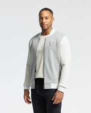 A person stands confidently in the MENS HYDRA COLOR BLOCK KNIT BOMBER - B6J455F200 by Psycho Bunny. The light gray varsity jacket, crafted from a premium cotton-blend with white accents and a white shirt, features an embroidered Bunny logo on the chest against a plain white background.
