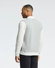 A man faces away, wearing the Psycho Bunny MENS HYDRA COLOR BLOCK KNIT BOMBER - B6J455F200, a gray and white varsity jacket made from premium cotton-blend. Light sleeves enhance its subtle design, featuring an embroidered Bunny detail. He glances sideward against a plain background.