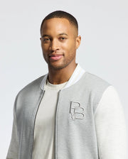 Someone is wearing a Psycho Bunny MENS HYDRA COLOR BLOCK KNIT BOMBER - B6J455F200 in light gray and white with an embroidered Bunny logo over a premium cotton-blend T-shirt. The background is plain white.