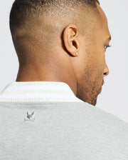 A man with short hair and a beard is seen from behind, wearing the Psycho Bunny MENS HYDRA COLOR BLOCK KNIT BOMBER - B6J455F200, featuring a light gray premium cotton-blend and white collar with an embroidered Bunny logo. A stud earring glints in his left ear.