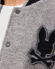 Close-up of the Psycho Bunny MENS TOM WOOL BOMBER JACKET - B6J744D200 in gray, featuring a black and gray embroidered patch with a rabbit head design and crossed bones beneath it. The premium fabric has a textured appearance, with part of a white shirt visible at the neck.