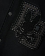 Close-up of the Psycho Bunny Men's Brayden Wool Leather Varsity Bomber - B6J876D200 in black, featuring leather sleeves and a prominent patch. The patch showcases a stylized rabbit head above crossbones, positioned against two large interlocking "BB" letters crafted from textured fabric. This wool-blend jacket is finished with metal snap buttons.