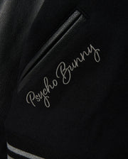 Detailed view of the MENS BRAYDEN WOOL LEATHER VARSITY BOMBER jacket, featuring "Psycho Bunny" elegantly embroidered in white cursive. This black bomber includes leather sleeves, a leather accent, and striped ribbed trim.