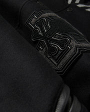 A detailed close-up of the MENS BRAYDEN WOOL LEATHER VARSITY BOMBER - B6J876D200 by Psycho Bunny showcases a black wool-blend fabric with an intricate embroidered design featuring crossbones and decorative elements. The raised and detailed stitching adds texture against the smooth background, reflecting the exceptional craftsmanship typical of a varsity bomber.