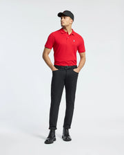 In a plain white background, a man stands with hands in pockets wearing a Psycho Bunny MENS CLASSIC PIQUE POLO SHIRT - B6K001CRPC in red, paired with a black cap, black pants, and black boots.