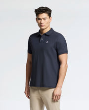 A man in a Psycho Bunny Men's Classic Pique Polo Shirt (B6K001ARPC) in navy, featuring 100% Pima cotton, an embroidered logo, and mother-of-pearl buttons, pairs it with beige pants against a plain light background.