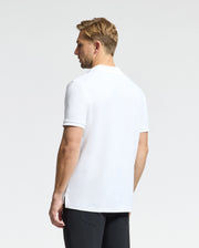 A man with short blonde hair, dressed in a Psycho Bunny MENS CLASSIC PIQUE POLO SHIRT - B6K001ARPC made of soft Pima cotton, is wearing black pants. The white polo shirt displays the Psycho Bunny logo while he stands against a plain light gray background, gazing slightly to the right.
