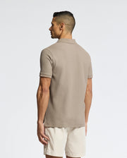 A man faces away, wearing the Psycho Bunny MENS CLASSIC PIQUE POLO SHIRT - B6K001CRPC in taupe Pima cotton, paired with light beige shorts, set against a plain light background.