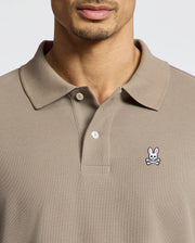 A man wears a taupe Psycho Bunny MENS CLASSIC PIQUE POLO SHIRT (B6K001CRPC) with a collar and buttons, made from soft Pima cotton. A pink bunny and white skull-and-crossbones patch is on the left chest. His face is partially visible.