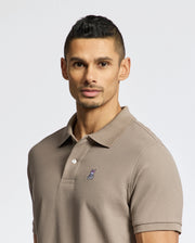 A man with short dark hair and light facial hair wears a Psycho Bunny MENS CLASSIC PIQUE POLO SHIRT - B6K001CRPC in taupe, featuring a small embroidered bunny head on the chest, standing against a plain light background.