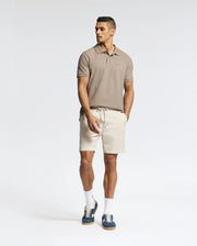 A man stands against a white background wearing a Psycho Bunny MENS CLASSIC PIQUE POLO SHIRT (B6K001CRPC) in beige, paired with light beige shorts, white socks, and navy sneakers. His hands are in his pockets as he looks to the side.