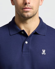 A man is dressed in a regular fit, navy blue polo shirt made from soft pima cotton, displaying the pink Psycho Bunny logo with crossbones on the chest. The backdrop is simple and light-toned.