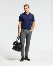 A man wearing a Psycho Bunny MENS CLASSIC PIQUE POLO SHIRT in navy and gray trousers stands against a plain backdrop. He holds a black duffel bag in one hand, complemented by black shoes and a watch.