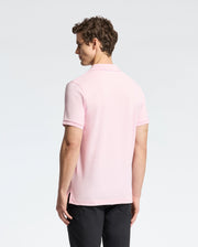 A curly-haired man is seen from behind, donning the Psycho Bunny light pink MENS CLASSIC PIQUE POLO SHIRT - B6K001CRPC in luxurious pima cotton with black pants, against a plain light backdrop.