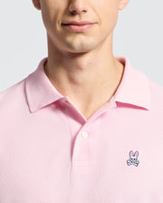 A person is wearing the Psycho Bunny MENS CLASSIC PIQUE POLO SHIRT - B6K001CRPC in pink, featuring a bunny and crossbones logo on the left chest. The photo highlights their upper chest and partially visible face against a simple light background.