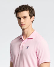 A person with curly hair wears a Psycho Bunny MENS CLASSIC PIQUE POLO SHIRT - B6K001CRPC in light pink, made of luxurious Peruvian pima cotton, featuring a small embroidered bunny logo. They stand against a plain gray background, looking slightly to the side.