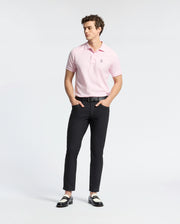 A person wearing a light pink Psycho Bunny MENS CLASSIC PIQUE POLO SHIRT - B6K001CRPC, black pants, and black and white loafers stands against a plain white background. They have curly hair and their hands are in their pockets.