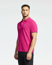 A man poses against a plain background in Psycho Bunny's bright pink MENS CLASSIC PIQUE POLO SHIRT - B6K001CRPC with an embroidered Bunny logo, paired with black pants. His short curly hair and neatly trimmed beard enhance his casual yet confident look.