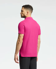 A man with short curly hair faces away from the camera wearing a Psycho Bunny MENS CLASSIC PIQUE POLO SHIRT - B6K001CRPC in bright pink. Made of soft Pima cotton and featuring an embroidered Bunny logo, it pairs perfectly with his black pants against a light gray background.