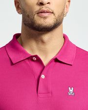 A person is wearing a Psycho Bunny Men's Classic Piqué Polo Shirt (B6K001CRPC) in bright pink, featuring an embroidered Bunny logo with crossbones on the chest. Made of soft Pima cotton, it includes a stylish collar and button details. The background is plain light gray.