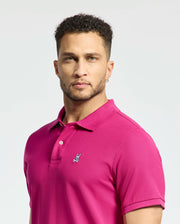 A person with short curly hair, wearing a Psycho Bunny MENS CLASSIC PIQUE POLO SHIRT - B6K001CRPC in bright pink, stands against a light gray background. They have a neutral expression and an earring in their left ear.