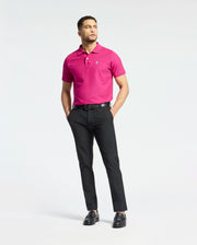 A man confidently poses against a white backdrop, wearing a bright pink Psycho Bunny Men's Classic Piqué Polo Shirt (B6K001CRPC) with an embroidered Bunny logo. He pairs it with black pants and shoes, hands in pockets, gazing sideways.