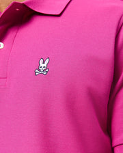 A close-up of a person wearing the Psycho Bunny MENS CLASSIC PIQUE POLO SHIRT - B6K001D200 in 100% Pima cotton shows a small embroidered chest logo with a white cartoon bunny and crossbones. Made in Peru, it features a buttoned collar.