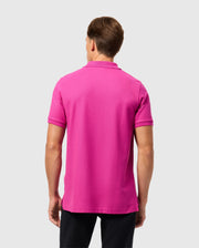 A person with short hair is wearing a Psycho Bunny MENS CLASSIC PIQUE POLO SHIRT - B6K001D200 in bright pink, made from 100% Pima cotton in Peru, paired with black pants. They stand back to the camera against a plain gray background.