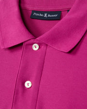Close-up of the Psycho Bunny Men's Classic Pique Polo Shirt, B6K001D200, in magenta, made in Peru from 100% Pima cotton. It features white buttons and a stitched "Psycho Bunny" label with a bunny logo inside.